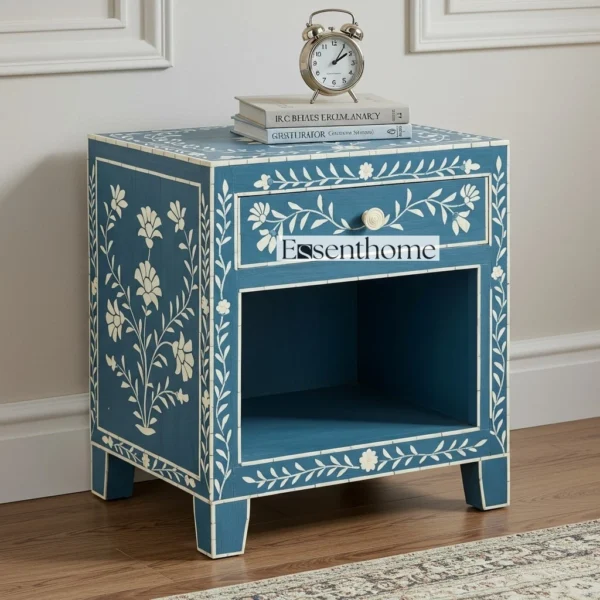 Handmade blue and white floral bone inlay bedside table with one drawer and an open shelf. Features intricate white inlay patterns on a deep blue background, a round bone knob, and short tapered legs. Styled with books and a clock on top, placed against a white paneled wall on a wooden floor.